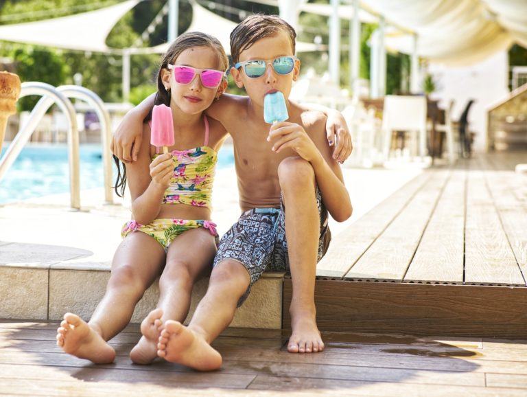 Portrait of little girl and boy with popsicles wearing mirrored sunglasses in front of swimming pool